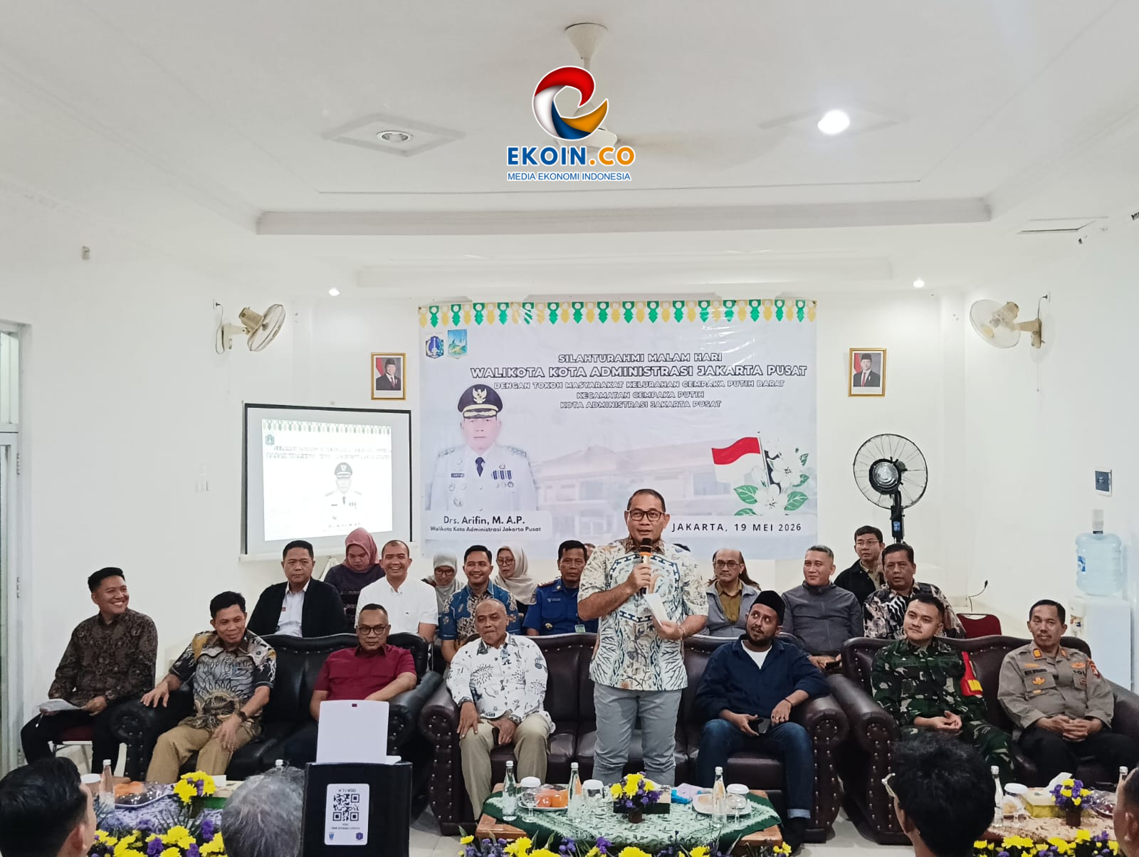 Speaker with microphone standing in a meeting room, banner and attendees seated on sofas behind him, Indonesian flags visible in the background