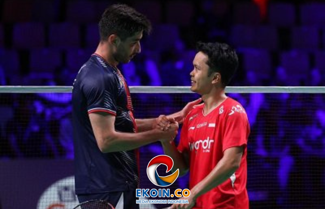 Two badminton players shake hands at the net after a match, one in a dark blue shirt and the other in a red jersey, in a dimly lit arena with a purple glow.