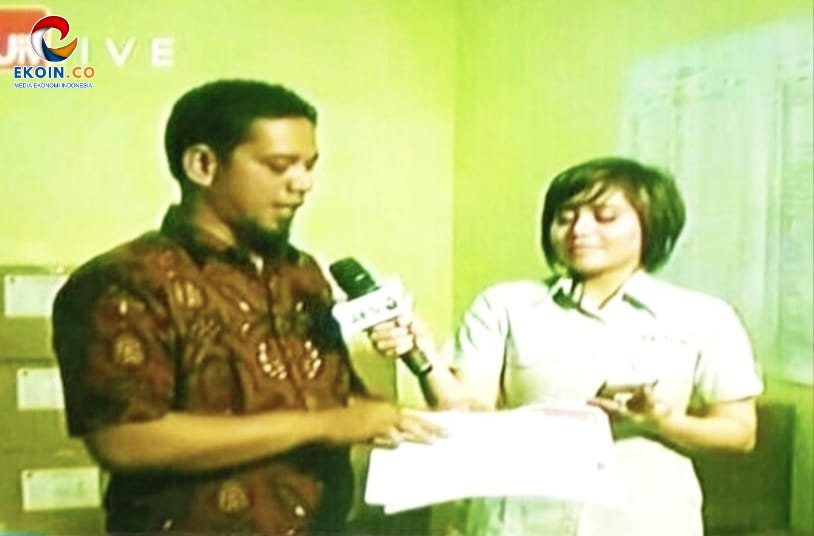 Reporter interviews a man indoors, microphone in hand while she holds a document; lime-green wall in the background.