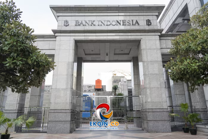 Stone entrance to Bank Indonesia building with a wide rectangular portal, flanked by trees; EKOIN.CO logo visible in the foreground.