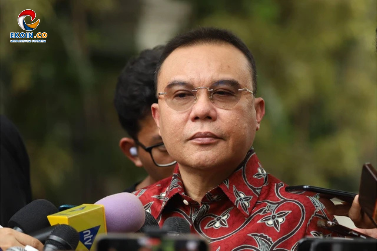 Man wearing glasses in a red batik shirt being interviewed by reporters outdoors, microphones in foreground, EKOIN logo top-left