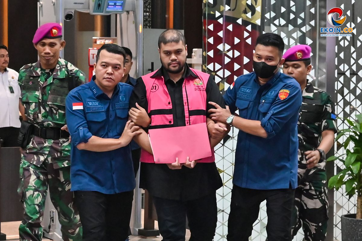 Man in a pink vest is escorted by two blue-uniform officers through a doorway, security personnel nearby in a building.