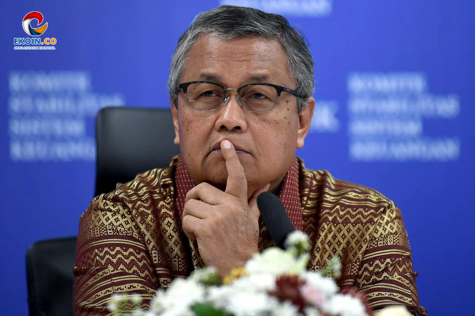 Man wearing glasses and a patterned batik shirt at a press conference, finger resting on his lips in a thoughtful pose by a microphone.