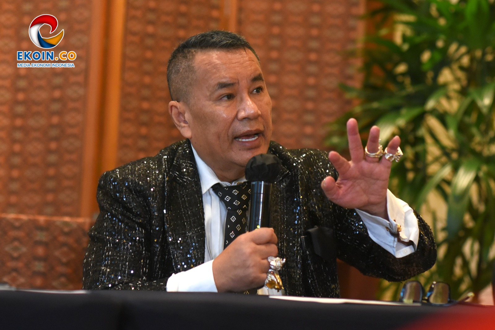 Speaker wearing a glittery black suit and tie, speaking into a handheld microphone with one hand raised, EKOIN.CO logo visible in the top-left corner.