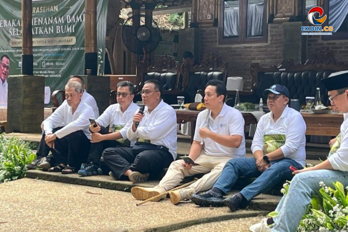 Group of men in white shirts sit on a low stage outdoors; one man speaks into a microphone while others listen. Banner and EKOIN.CO logo visible in background.