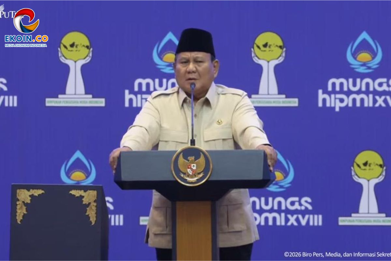 Indonesian official in beige uniform and black songkok giving a speech at a podium with national emblem, blue backdrop logos behind him.