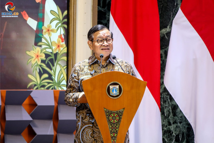 Smiling man in batik shirt delivering a speech at a wooden podium with a crest, microphones in front, flanked by Indonesian red-and-white flags.