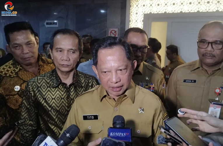 Central man in tan uniform being interviewed by reporters, microphones visible labeled Kompas TV and others, with colleagues around him in a dim indoor setting.