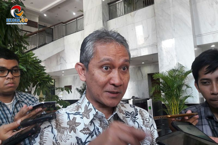 Middle-aged man in a floral shirt is interviewed by reporters in a marble lobby; journalists point recording devices toward him.