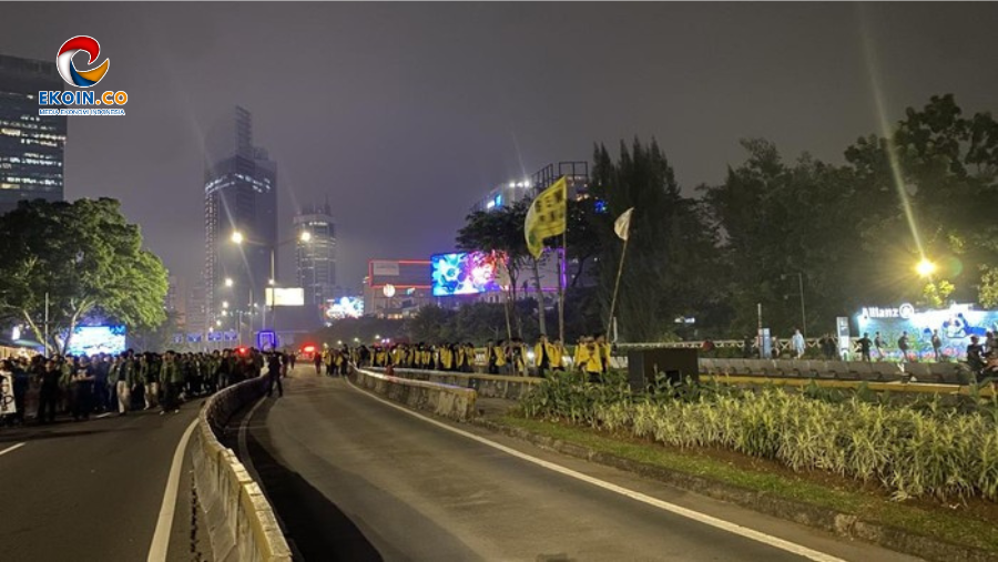 Nighttime street parade with a crowd along a curved divider, people in yellow vests marching, illuminated billboards and city buildings in the background.