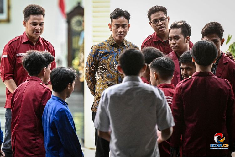 Group of young men in maroon uniforms listen to a man in a batik shirt in a bright indoor setting.