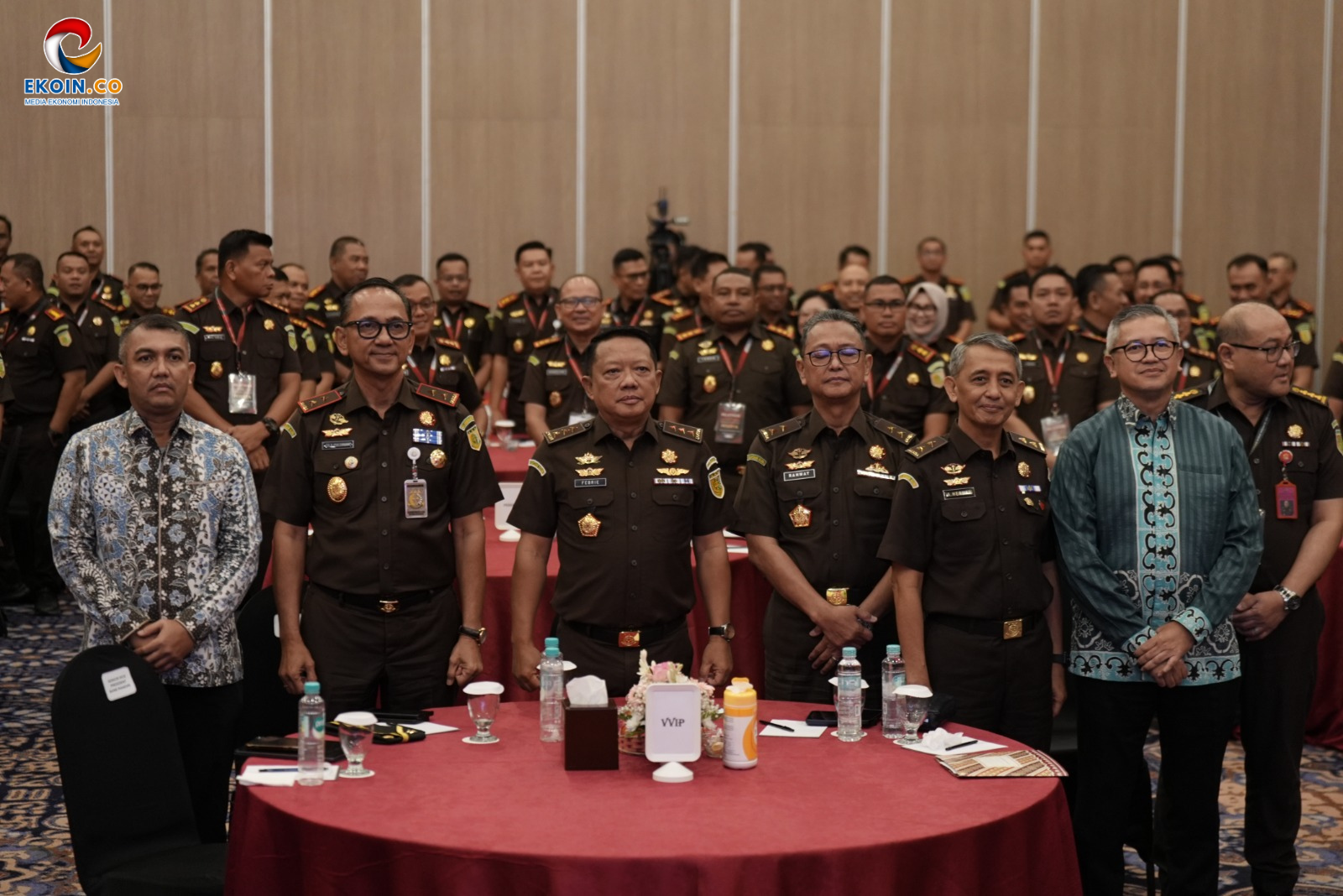 Group of uniformed officials standing around a round red-draped table in a formal event, with a VIP sign and water bottles visible in front. In the background, many attendees in uniform form a crowd.