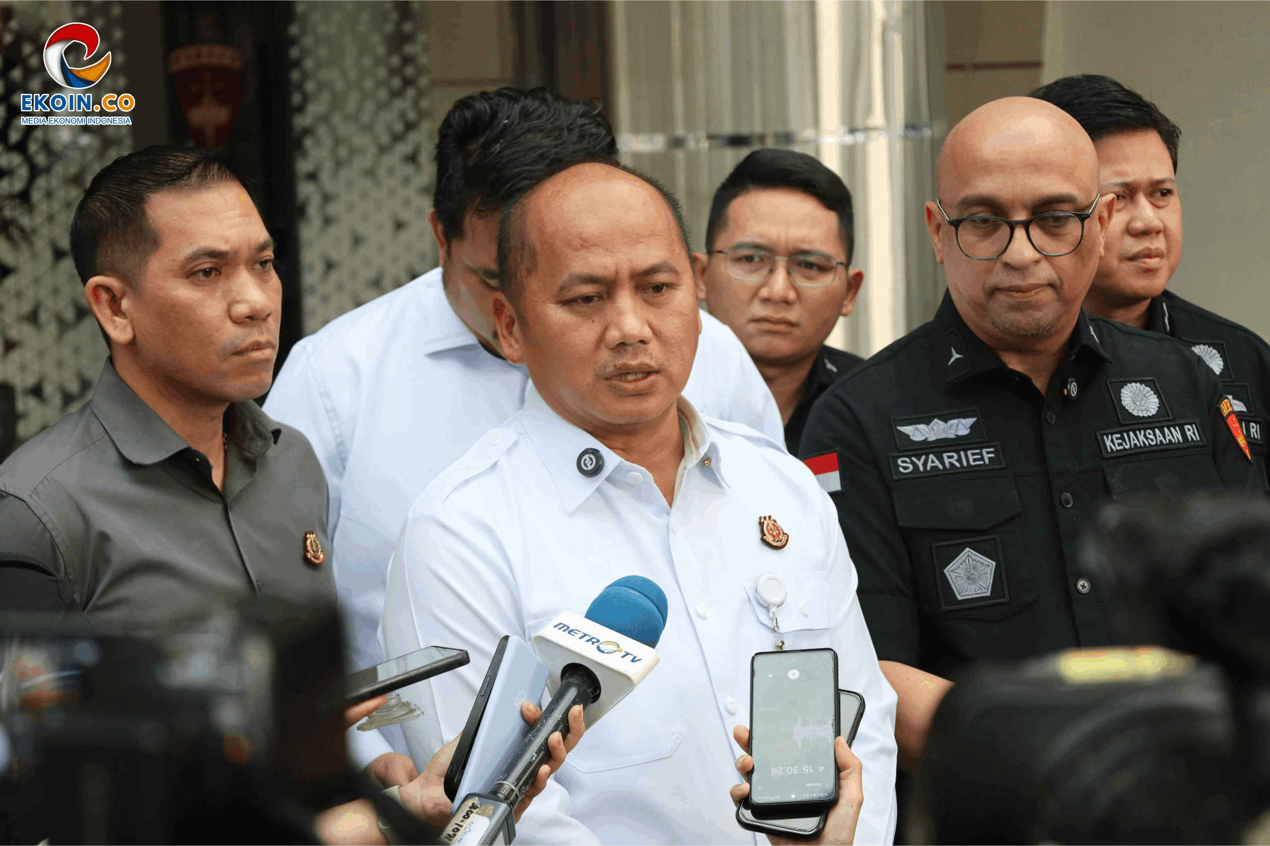 Officials speak to reporters at a press conference, microphones in the foreground and EKOIN.CO logo in the top-left corner
