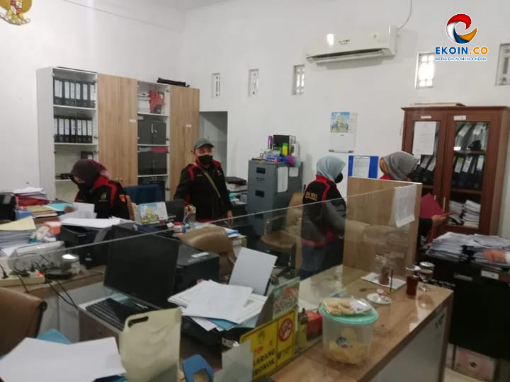 Office scene with several staff at desks, wearing masks and red-and-black jackets, working among papers and computers; glass divider on the counter and filing cabinets in the background.