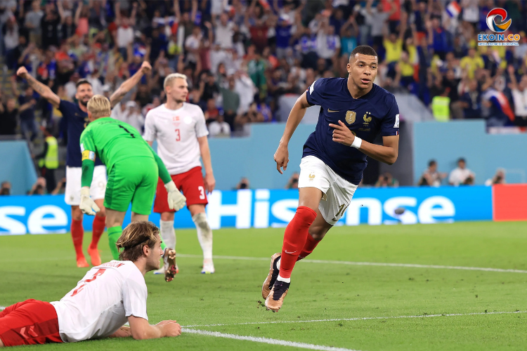 French soccer player in navy kit runs toward the goal as a sliding opponent in white challenges and a green-clad goalkeeper watches nearby.