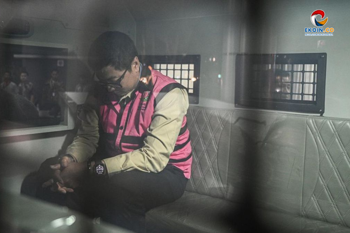 Man in a pink safety vest sits on a quilted gray seat inside a vehicle, hands clasped and head tilted down.