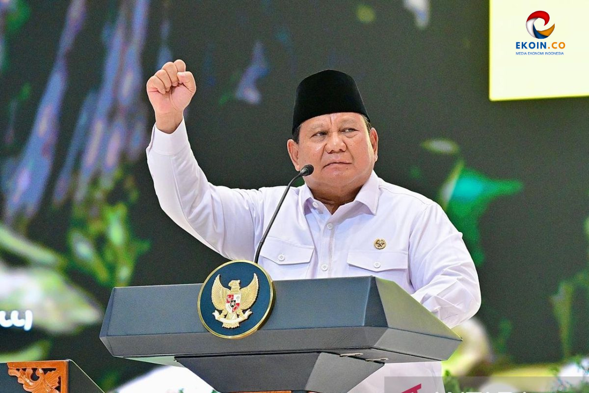 Male Indonesian official in white shirt and peci raises fist while speaking at a podium with Garuda emblem, EKOIN.CO logo visible in the corner