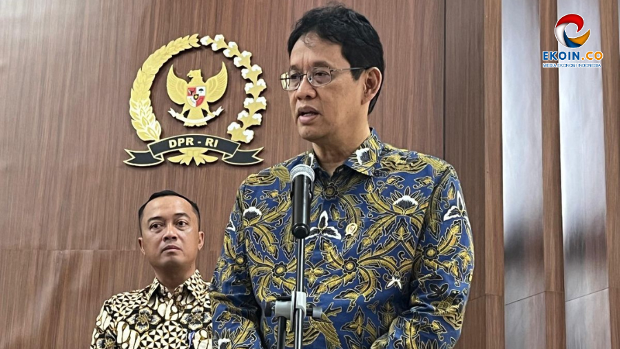 Man in a blue and gold batik shirt speaks at a microphone during a press briefing, with the DPR-RI emblem on a wooden wall behind him and another man in a patterned shirt in the background.