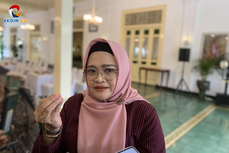 Woman in a pink hijab and glasses sits at an indoor event, gesturing with her hand as she talks or explains something. The EKOIN.CO logo is visible in the corner.