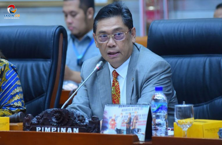 Man in a gray suit and red tie speaking into a microphone at a formal meeting, seated at a panel desk with a 'BIMPINAN' nameplate in front of him.