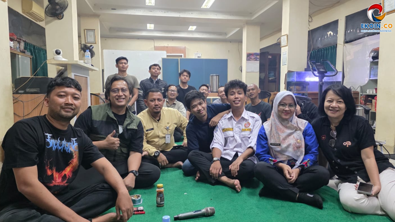 Group of adults sitting on a green carpet in a room, smiling at the camera during a casual gathering or meeting.
