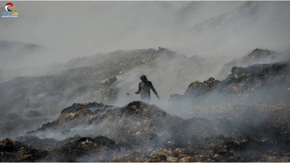 Person wearing protective gear walks through a smoky, rocky landfill terrain, holding a tool