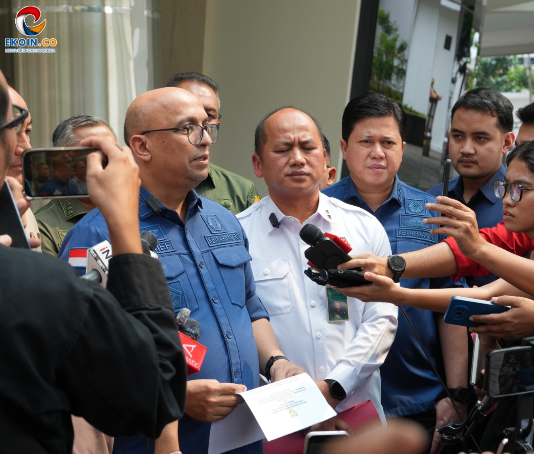 Officials in blue uniforms address the media during a press briefing outdoors, microphones held near them by reporters, logo visible in corner.