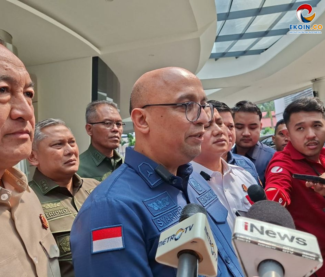 Indonesian police officer in a blue uniform speaks to reporters, with microphones from Metro TV and iNews nearby outside a curved building.