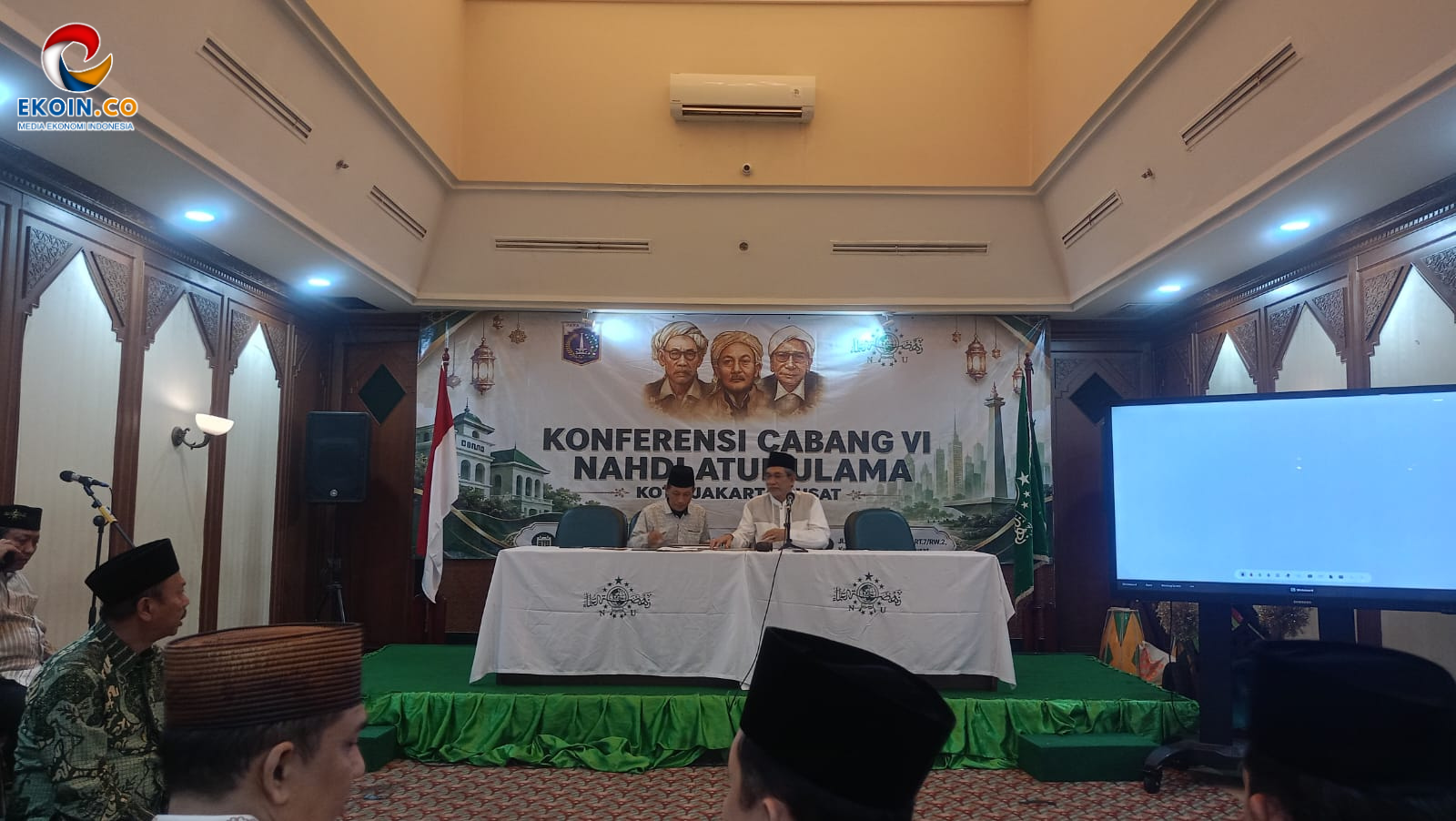 Panel of two men seated at a head table on a stage during a Nahdlatul Ulama conference, Indonesian flag on the left and a large screen on the right.
