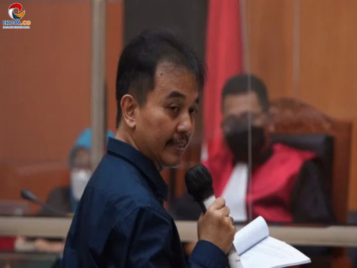 Man in a dark blue shirt speaks into a handheld microphone in a courtroom, papers in hand, with a judge seated behind glass.