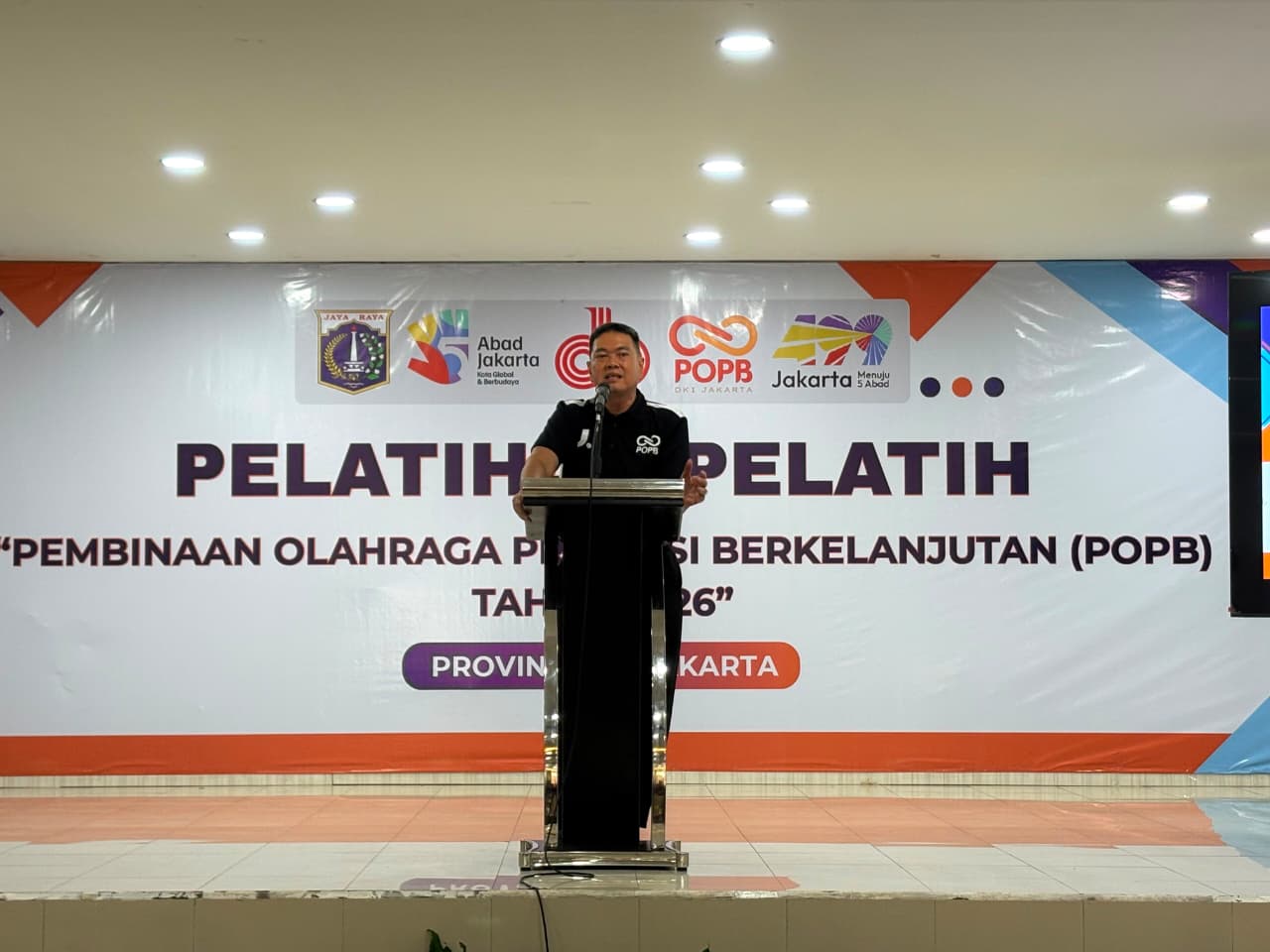Speaker at a podium giving a talk during a training event in front of a banner with PO DPB Jakarta logos and bold Indonesian text