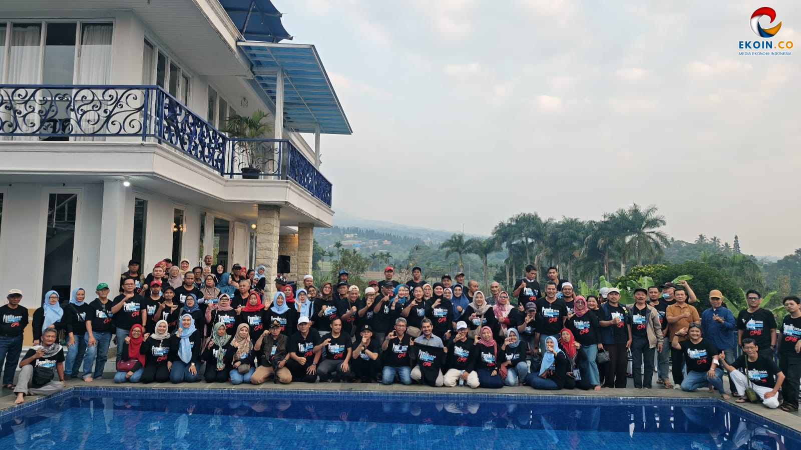 Group photo of dozens of people in matching black shirts posing by a pool at a resort with a white building and blue railings behind them.