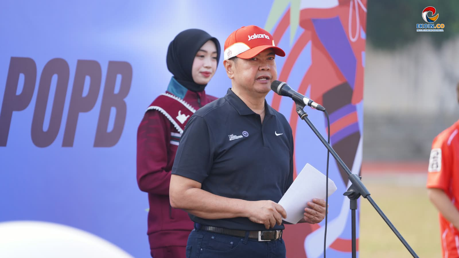 Man in a black polo and red Jakarta cap speaks into a microphone at an outdoor event, holding papers with a woman in a hijab behind him.
